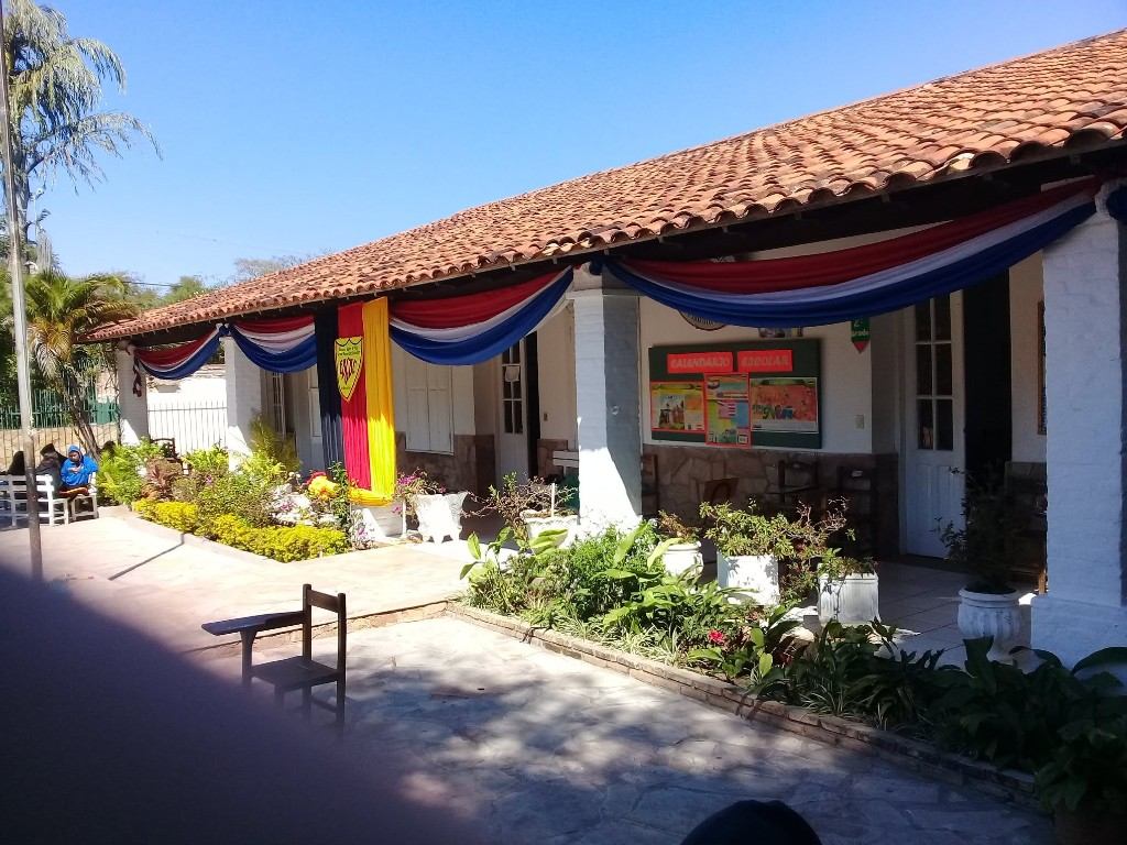 Patio interno de la Escuela Básica N° 1232 General Bernardino Caballero, en San Bernardino, con el cartel del calendario escolar y banderas de Paraguay en las columnas